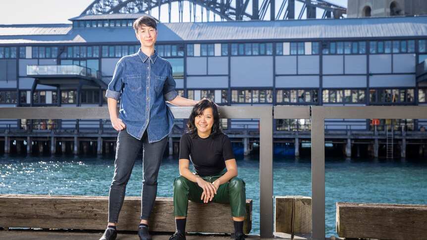 Two 30-something Asian Australian women, one standing, the other sitting, are by the harbour, the Harbour Bridge behind them