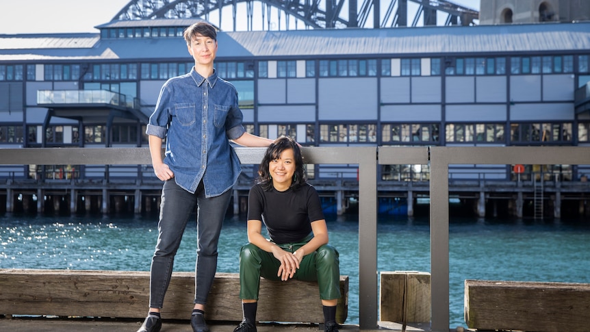 Two 30-something Asian Australian women, one standing, the other sitting, are by the harbour, the Harbour Bridge behind them