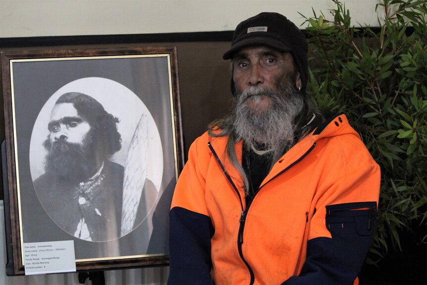 An Aboriginal elder sits next to the historic photograph of an Aboriginal man 