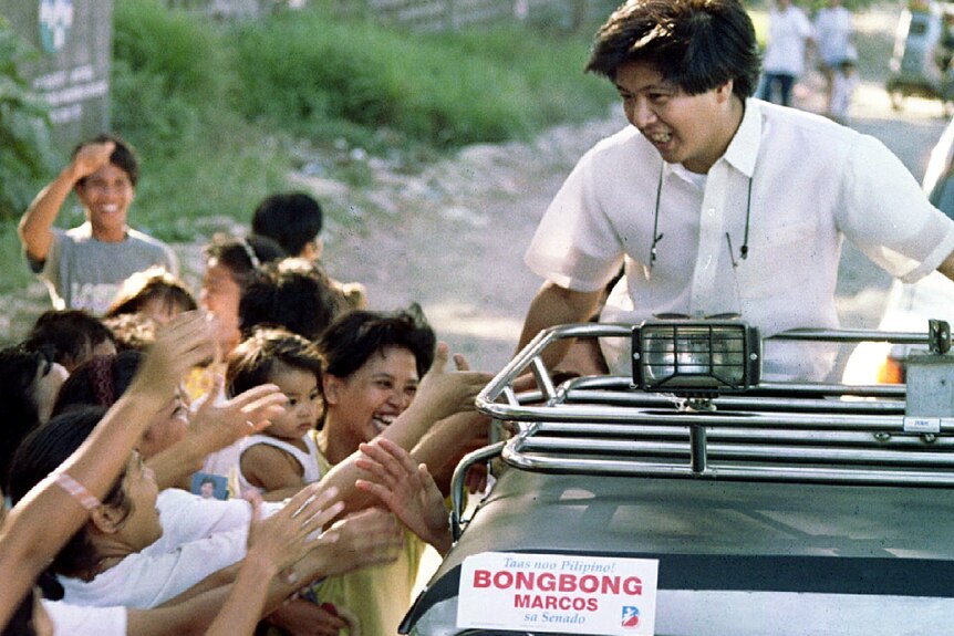 A young Filipino man rides on top of a car, greeting supporters