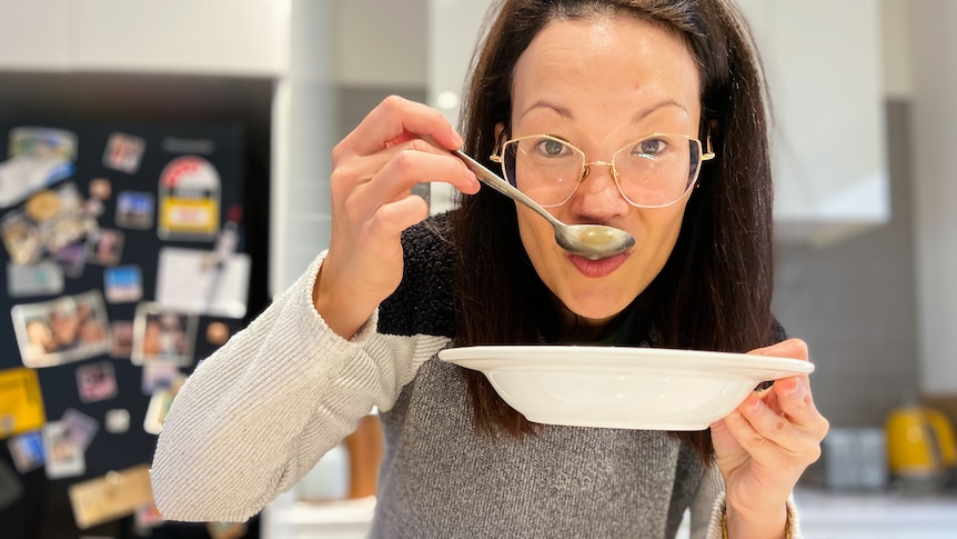 A woman holding a white bowl to her chin slurps soup from a spoon.