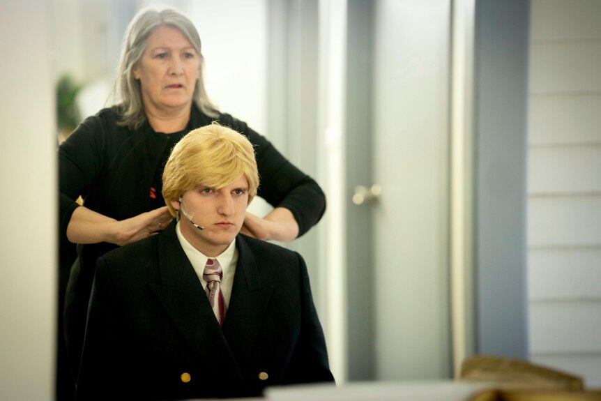 A teenage boy has a blonde wig fitted in a dressing room.