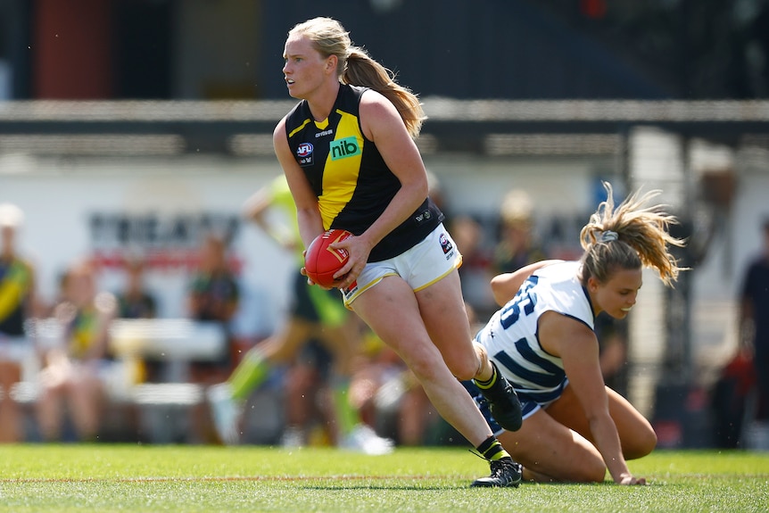 An AFLW player looks downfield as she holds the ball in her hands as a defender lies on the ground behind her..