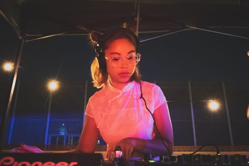 Maggie wears headphones as she works a DJ booth, hair in space buns.