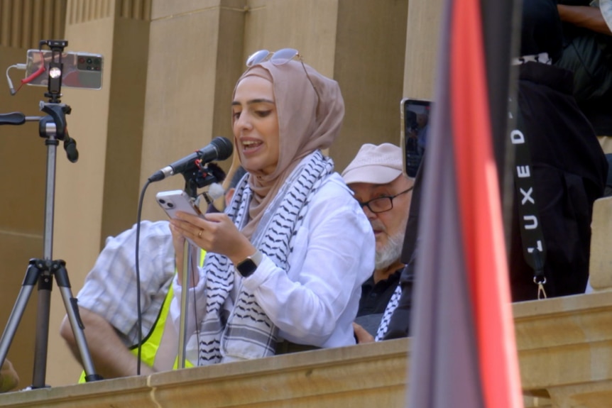 Woman wearing hijab and sunglasses on her head stands at lectern reading speech from mobile phone