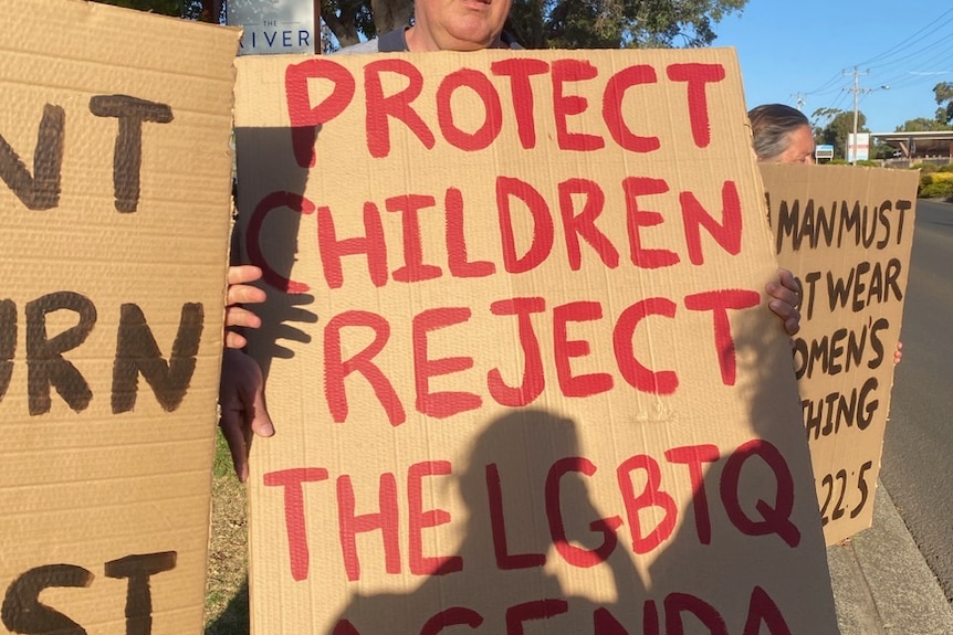 A man holds a cardboard sign with anti-LGBTQIA+ rhetoric painted on it in red