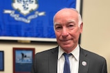 An older man with grey eyebrows wearing a blue tie and black suit jacket smiles in front of a blue Connecticut flag