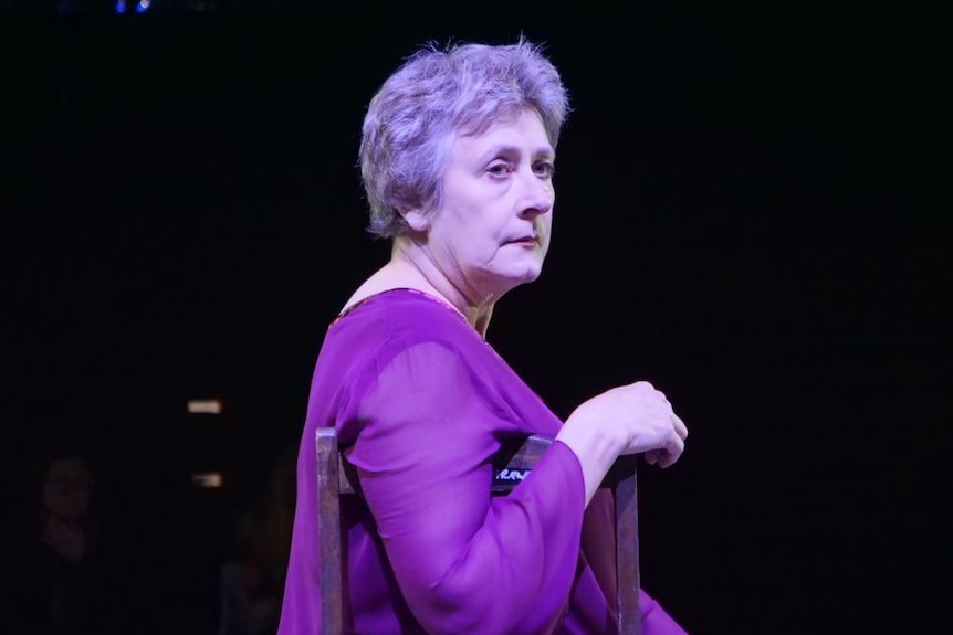 A female actor in a purple dress rests her arm on the back of a chair as she sits under a spotlight on stage.