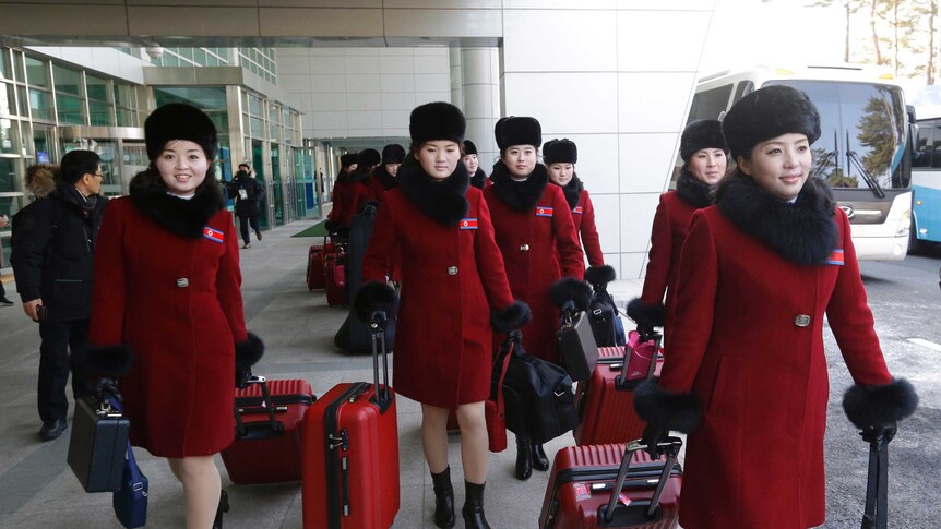 A group of North Korean cheering squad members in black-trimmed red coats walk through a transport terminal.