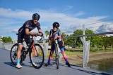 A father and his daughter on a bike wearing helmets and cycling gear smile at the camera.