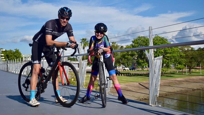 A father and his daughter on a bike wearing helmets and cycling gear smile at the camera.