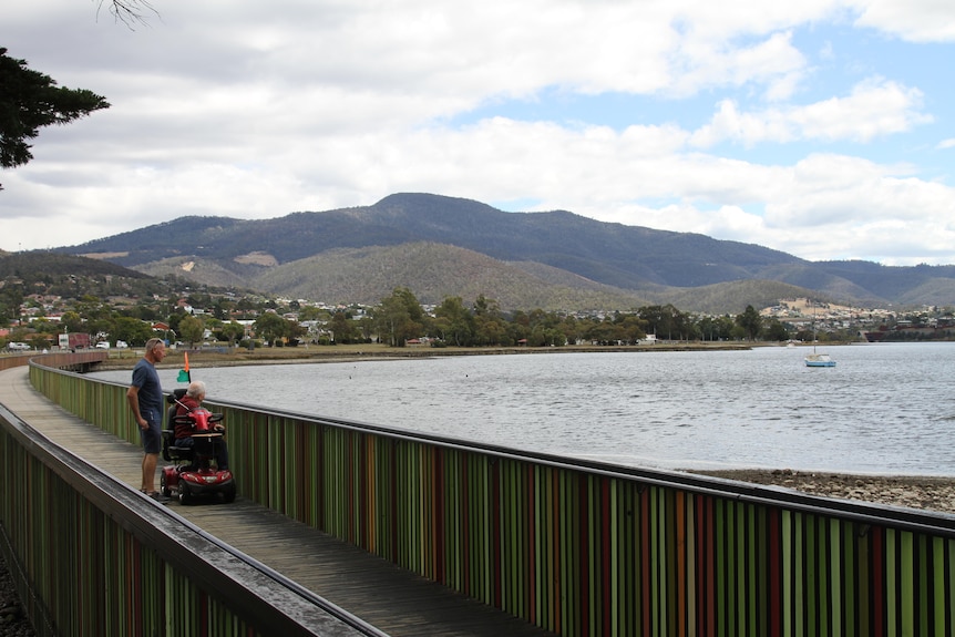 two men are on a boardwalk looking at the water. one is on a mobility scooter
