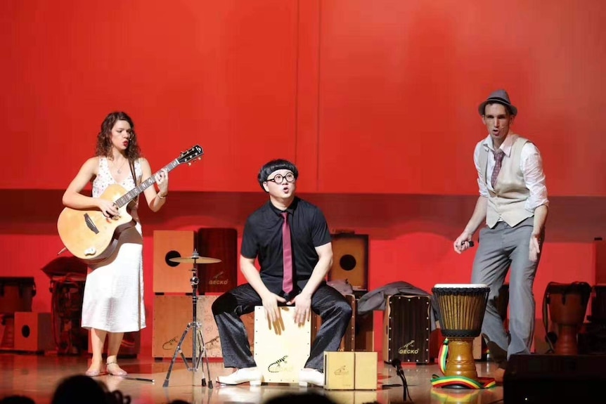 Australian musician Emily Pritchard holds a guitar jamming on the stage with other two performers with a red background.