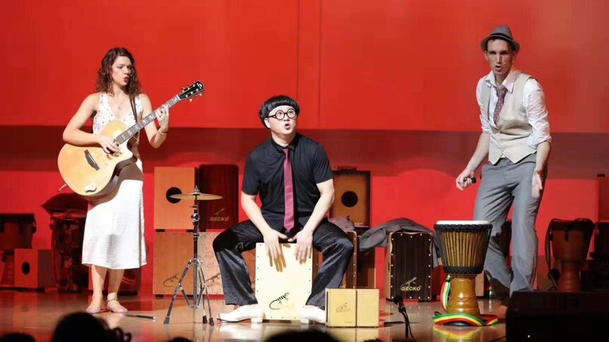 Australian musician Emily Pritchard holds a guitar jamming on the stage with other two performers with a red background.