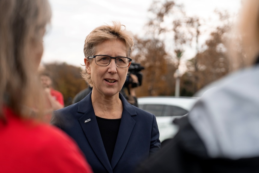 A woman with short light brown hair, glasses and a navy blazer speaking outside on an overcast day.