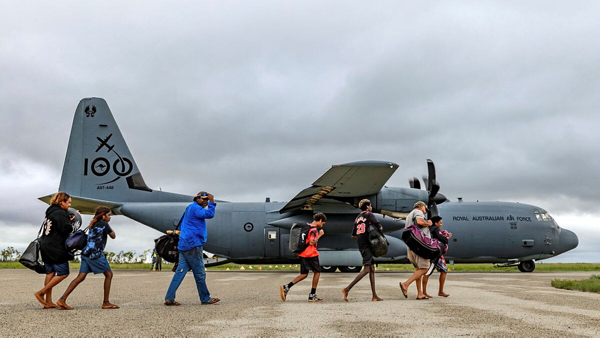 A line of people walk along the tarmac. A plane is in the background. 