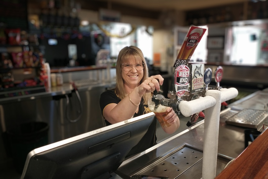 A blonde woman stands behind a bar pouring a glass of beer smiling at the camera 