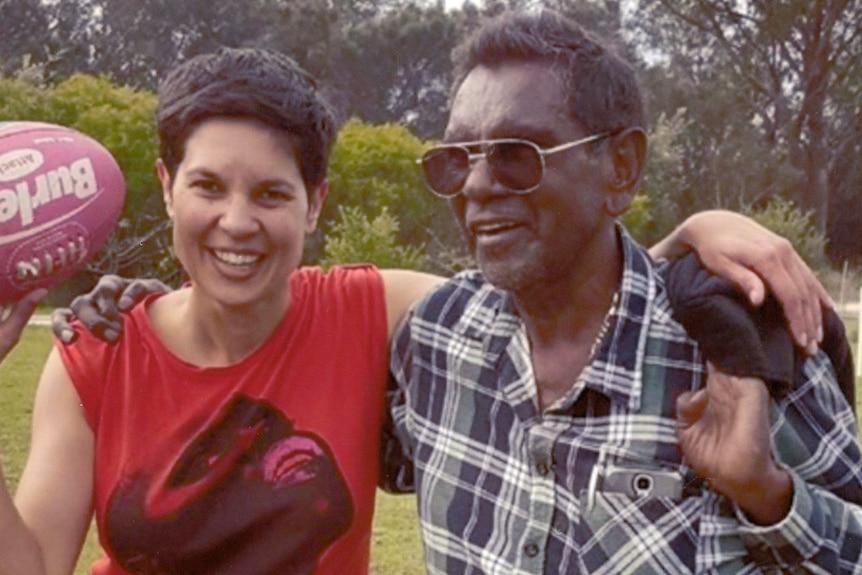 A photo of Narelda Jacobs in a red singlet holding a football with her father, who is wearing sunglasses and smiling