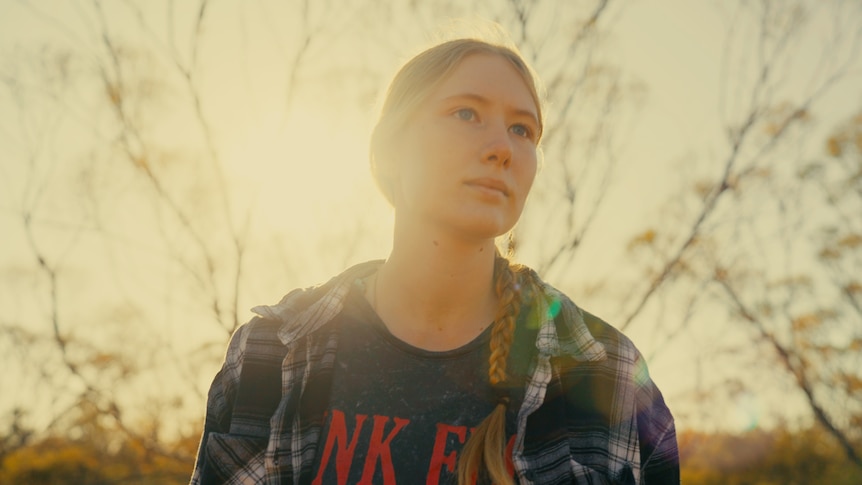 Outdoors, a young woman with blonde hair stairs into the distance, the sun glowing behind her head