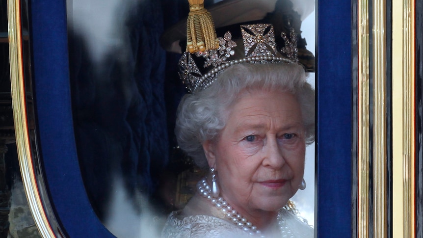 The Queen, wearing a crown and with a pensive expression, looks out of a horse-drawn carriage 