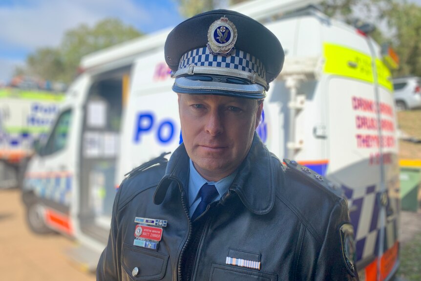 Head shot of a policeman wearing a hat and jacket in front of a police rescue van.