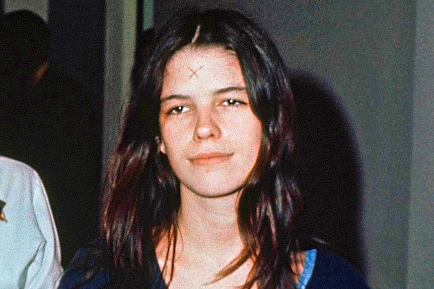 Leslie Van Houten looks past camera with vacant expression in grainy image. Her dark hair is down and a cross is on forehead.