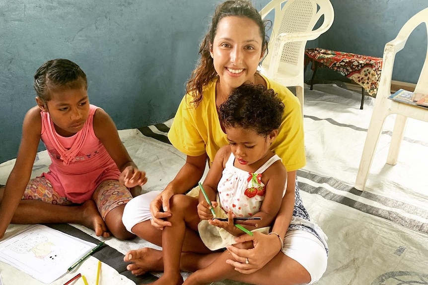 A smiling young woman with dark hair cradles a child in her lap. Another sits beside them.