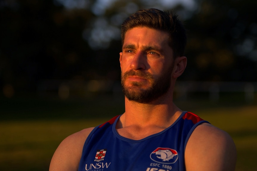 A man sitting on a bench looks ahead at the sunset with a neutral expression. He's wearing an Australian Rules jersey.