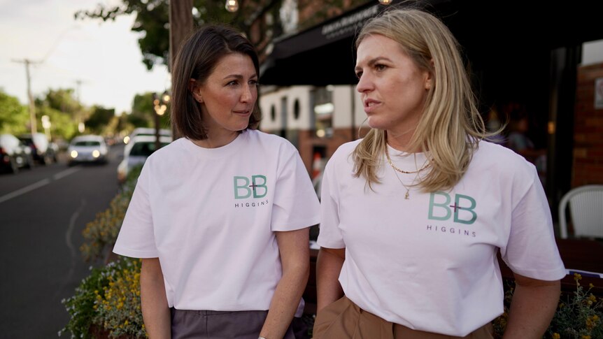 Two white women wearing white political campaign t shirts sit by the side of a road
