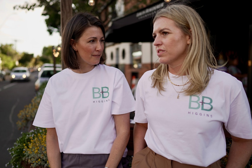Two white women wearing white political campaign t shirts sit by the side of a road