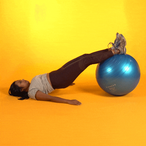 A young woman is lying on the floor on her back. Her heels are on an exercise ball and she is rolling the ball back and forth.