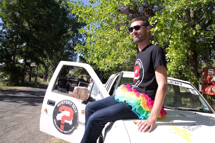 A man in a colourful tutu sits on a car with stickers on it that say mystery box
