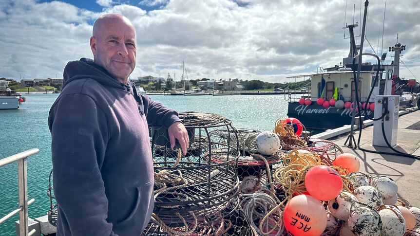 A man in a blue hoodie stands next to his lobster pots and buoys at a marina.