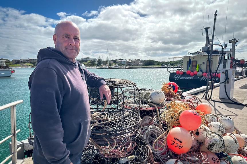 A man in a blue hoodie stands next to his lobster pots and buoys in at a marina. 
