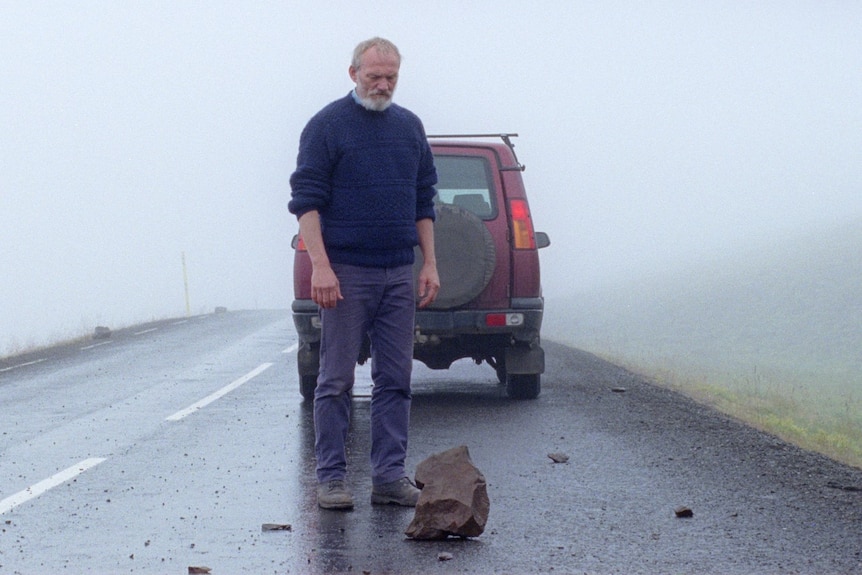 An older man standing on a road looking at a rock, car in the background, in the film A White, White Day