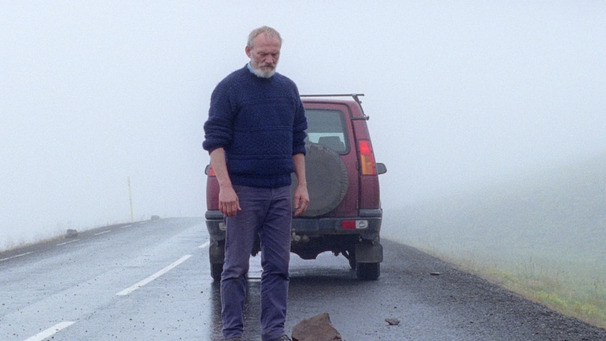 An older man standing on a road looking at a rock, car in the background, in the film A White, White Day