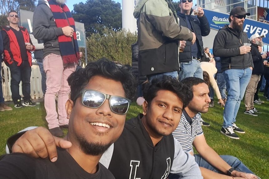 Three men take a selfie while sitting on the grass among other people standing up. 