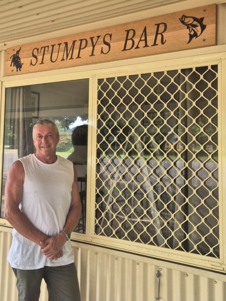 Man wearing white singlet standing in front of window with 'Stumpy's Bar' sign above
