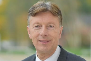 A man in a grey jacket with striped tie smiles slightly before a blurred background.