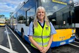 A blonde woman wearing a BCC branded hi-vis vest and large sunglasses smiles while staning in front of a bus. 