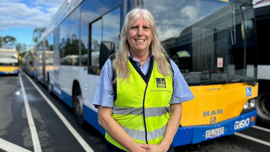 A blonde woman wearing a BCC branded hi-vis vest and large sunglasses smiles while staning in front of a bus. 