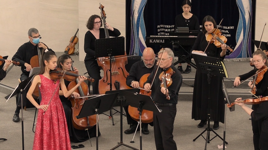 Ellie Malonzo plays violin in a red dress next to Paul Wright on violin in front of a small chamber orchestra