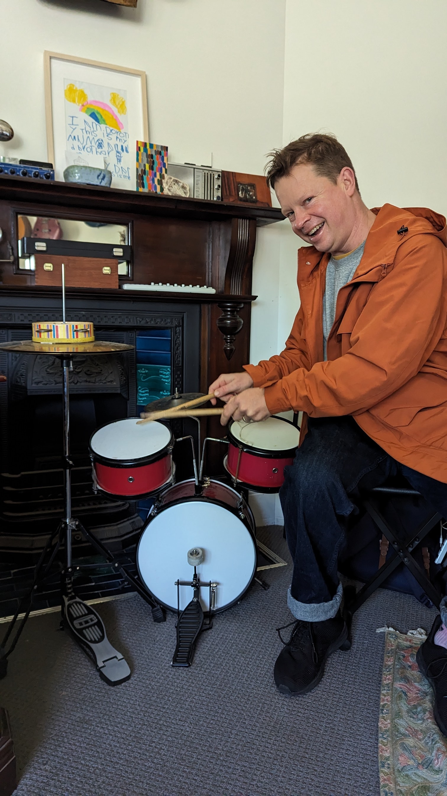 David wearing an orange jacket, sitting at a small drumkit playing it.