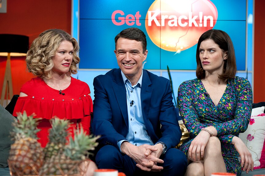 Two women with serious expressions, sit on either side of smiling man on couch on the set of a breakfast television show.