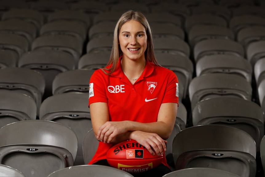 AFLW player sitting in the stands arms crossed atop a football. She is smiling directly at the camera.