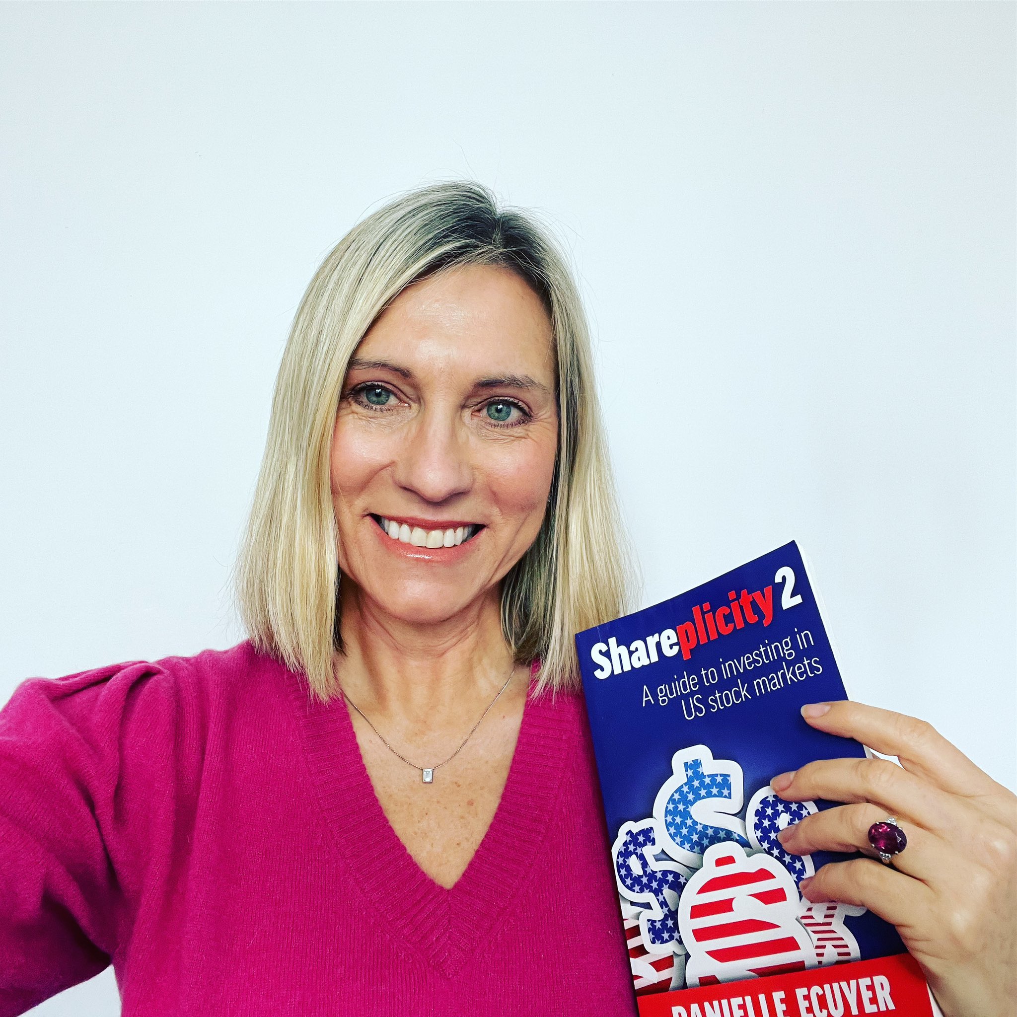 A woman with short blonde hair in a pink top poses for a selfie with a book