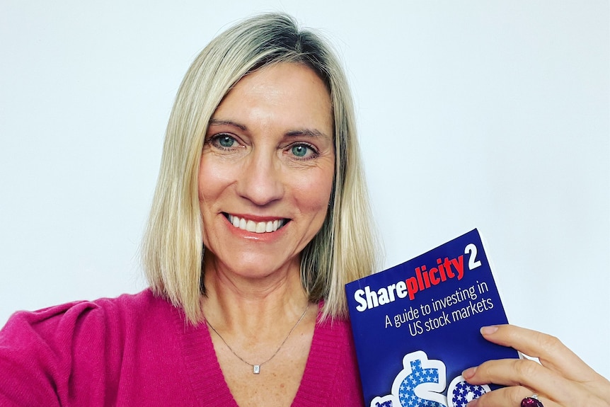 A woman with short blonde hair in a pink top poses for a selfie with a book