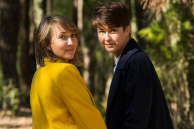 One woman in a bright yellow coat and one in a long black coat hold hands.