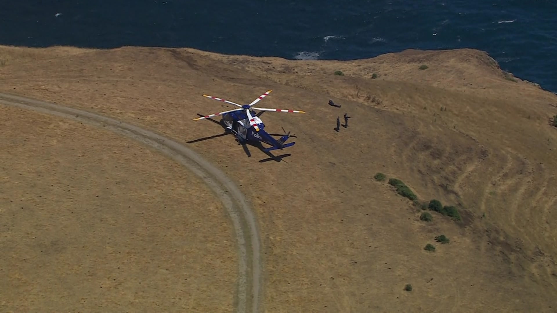 Un helicóptero azul y blanco se encuentra en la hierba de un acantilado junto al mar, con agua debajo.