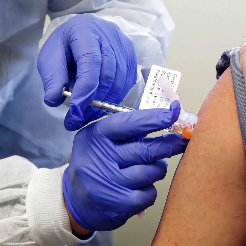 A pair of hands in latex gloves holds a vaccine vial and syringe as it is injected into an arm